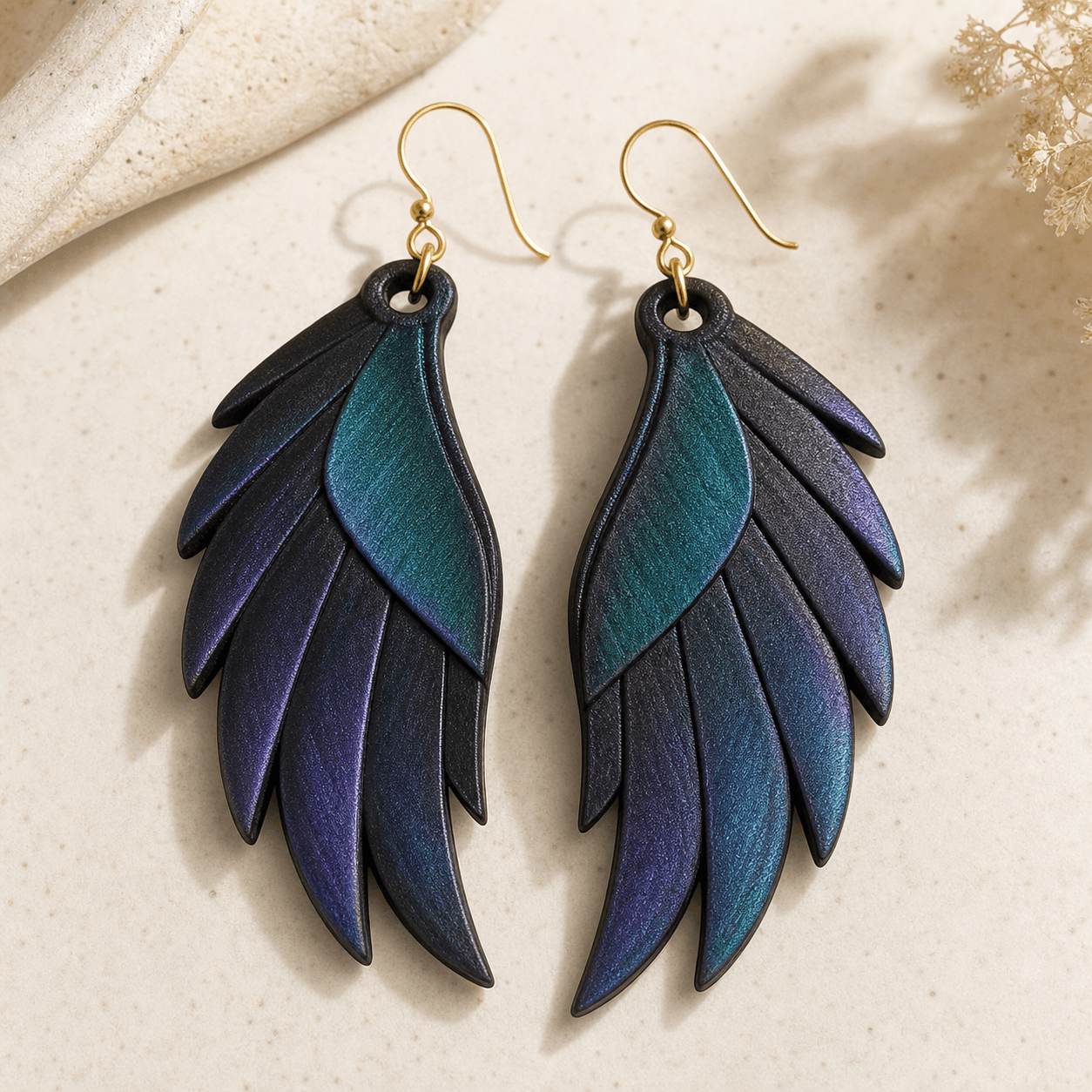 Grackle Wing Earrings styled flat on a stone surface with dried flowers, showing the layered feather detail